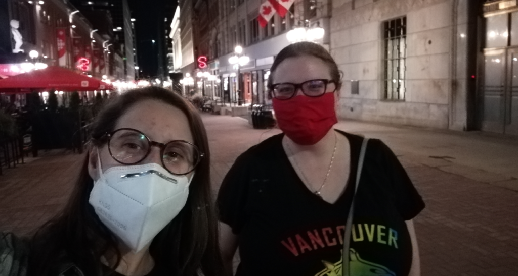 Taylor Fitpatrick y Ana Compañy en una calle peatonal. Hay banderas de Canadá de fondo. Ambas llevan mascarilla. Fitzpatrick lleva una camiseta de los Vancouver Canucks en colores arcoiris. 