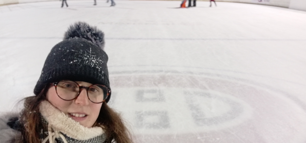 Selfie de una chica con gafas redondas y gorro de nieve. Se puede ver que está en una pista de hielo, hay gente patinando al fondo y se ve el logo de los Montreal Canadiens en el hielo.