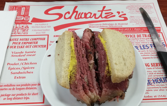 Un bocadillo partido en dos de carne ahumada con mostaza. Está sobre un mantel blanco en el que pone Schwartz's con letras rojas y hay información sobre los servicios del restaurante.