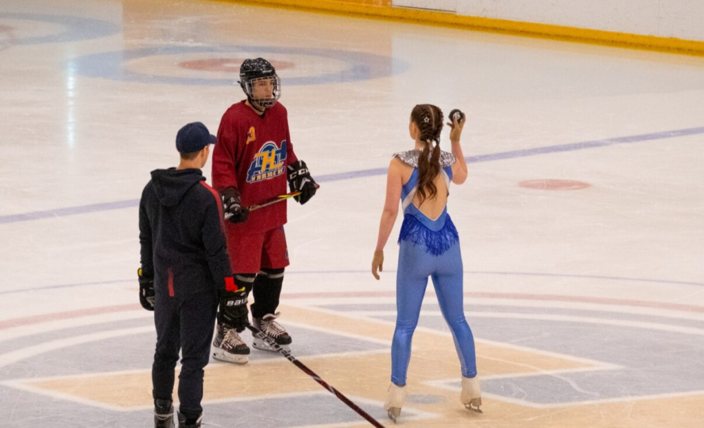 Kayla y Mac en el centro del hielo. Él lleva la equipación de hockey y ella un maillot de patinaje. Aparece también el entrenador del equipo, de espaldas.